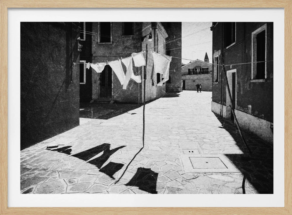 A framed, black and white photograph capturing a sunlit European alleyway. In the foreground, white laundry hangs on a clothesline, casting long, dramatic shadows across the cobblestone pavement. Old buildings line the alley, and in the distance, two people are walking away. Decor