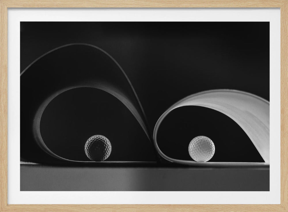 A framed, black and white abstract photograph featuring two golf balls nestled within the graceful curves of a single sheet of paper, creating a striking interplay of light, shadow, and form against a dark background. Decor