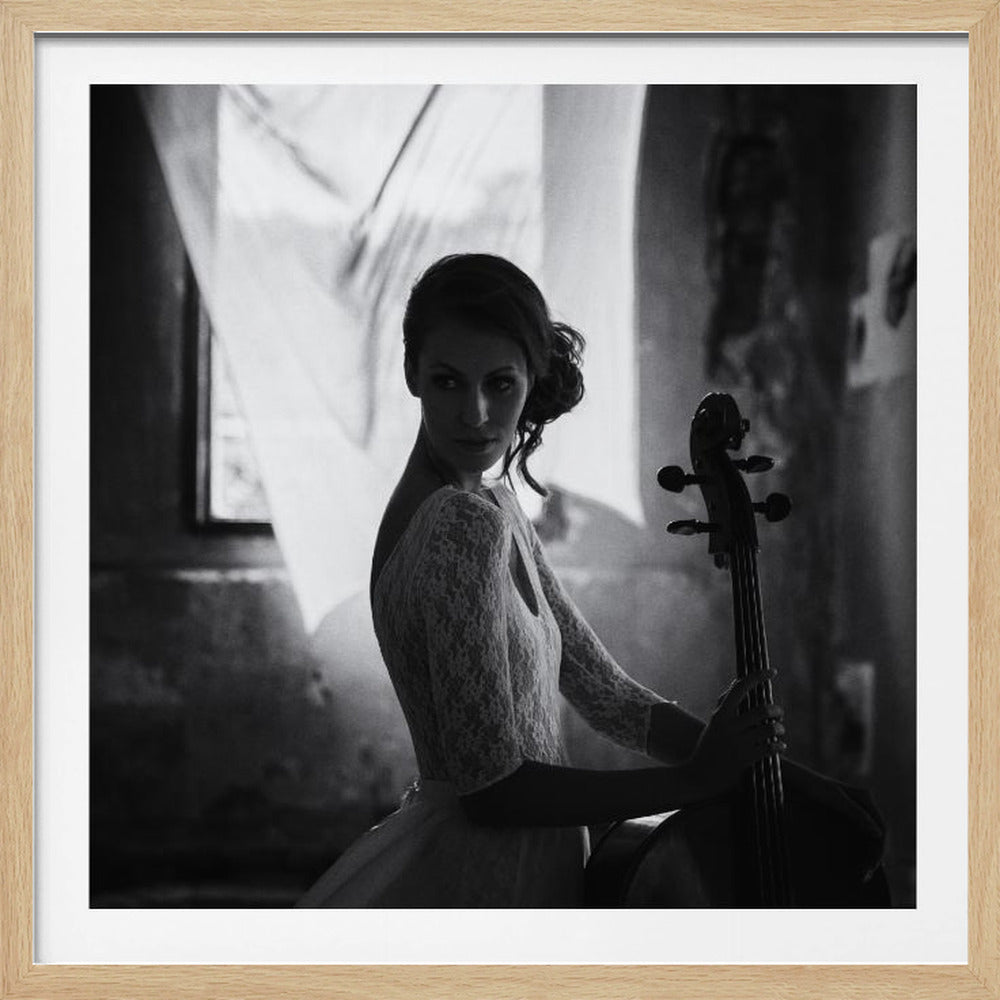 A framed, black and white photograph of an elegant woman in a lace dress holding a cello. She is looking over her shoulder directly at the camera with a subtle intensity. The background is softly lit, suggesting a room with a window and a sheer curtain. Wall Art