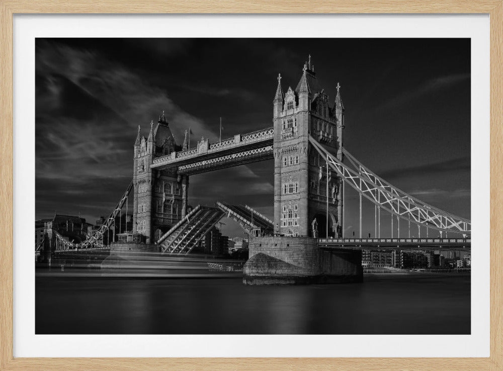 A dramatic black and white long-exposure photograph of London's Tower Bridge with its bascules raised to allow a ship, seen as a motion blur, to pass underneath on the River Thames. The sky is dark and cloudy, and the entire scene is enclosed in a silver frame. Artwork