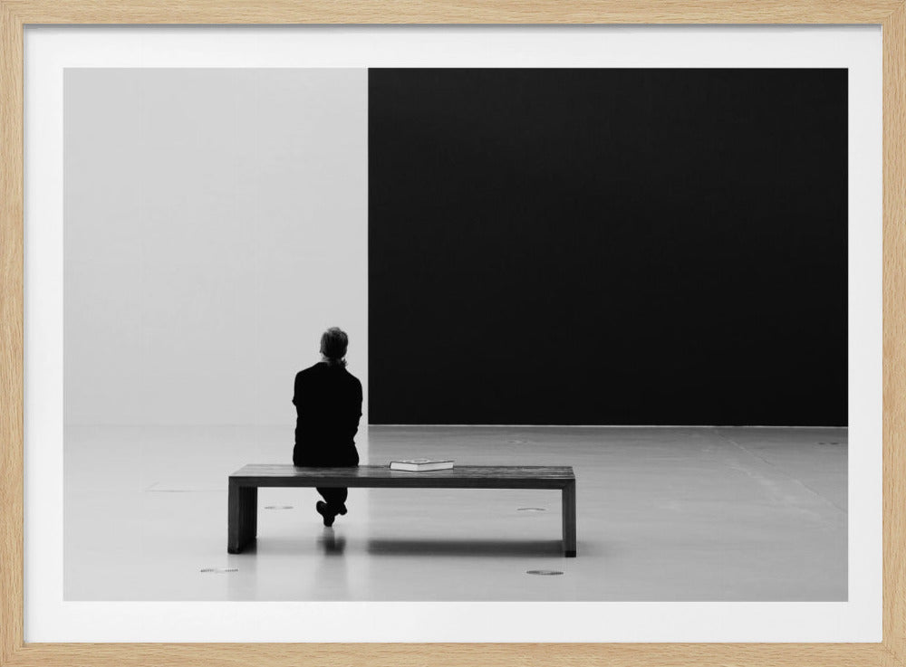 A black and white photograph of a person seen from behind, sitting alone on a wooden bench in a minimalist art gallery. They face a large, starkly divided wall, half white and half black. The scene is presented within a silver picture frame. Poster