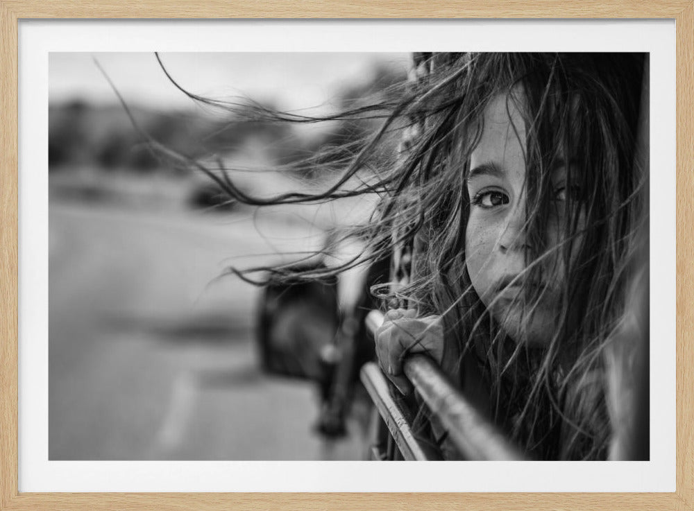 A close-up, black and white photograph of a young girl looking out of a moving vehicle. Her long hair is blowing wildly in the wind, partially obscuring her face as she peers intently at the camera with one eye. Her hand grips the edge of the open window. The background is blurred with motion, suggesting a journey. The photo is displayed in a silver frame. Poster
