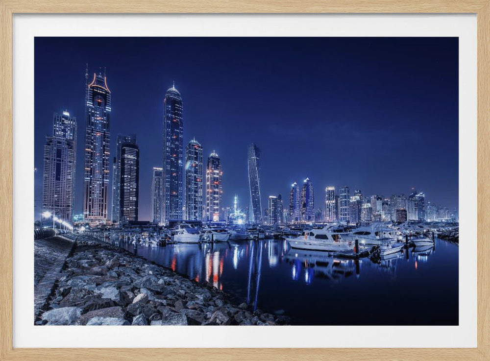 A stunning nighttime photograph of the Dubai Marina skyline, with illuminated skyscrapers towering against a deep blue sky. In the foreground, luxury yachts are moored in the calm harbor, their white hulls and the city's lights creating vibrant reflections on the water's surface. The shot is taken from a rocky embankment. Decor