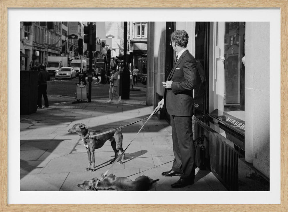 A black and white street photograph of a well-dressed man in a suit standing on a city sidewalk outside a storefront. He holds the leashes for two dogs; one is standing alert, and the other is lying on its back on the pavement. The background is a busy city street with traffic and pedestrians. Print