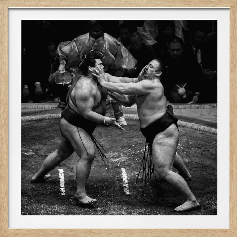A dynamic black and white action photograph of a sumo wrestling match. Two large wrestlers are in the middle of a clay ring, one pushing forcefully against the other's face. A referee in traditional robes stands behind them, and spectators watch from the background. The entire scene is captured within a light wood frame. Artwork