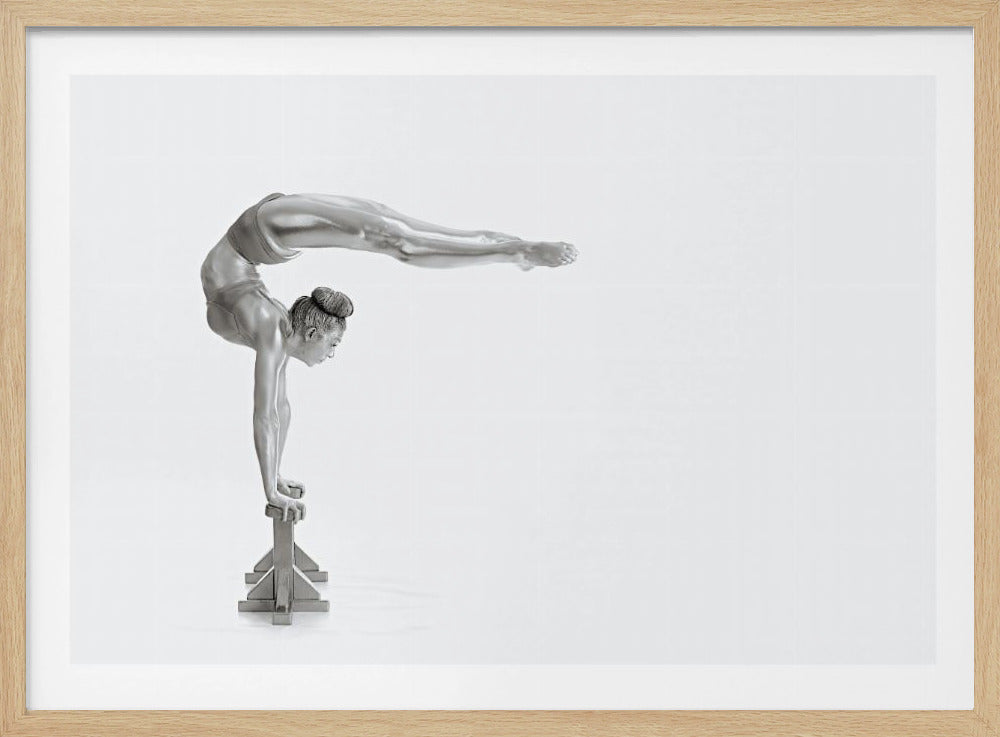 A framed black and white photograph of a female gymnast with a metallic silver-painted body, balancing in a difficult handstand pose on small blocks against a stark white background. Her back is arched and her legs are extended straight out behind her, parallel to the ground. Artwork