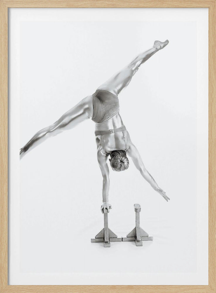 A black and white studio photograph of a muscular gymnast coated in silver paint, performing a dynamic one-armed handstand on a small block. Their legs are split wide apart in a display of strength and flexibility against a clean white background, all enclosed in a black frame. Decor