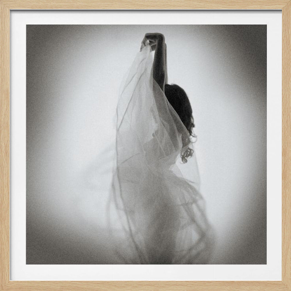 A framed, square black and white photograph of a woman seen from behind. She is holding a sheer, flowing veil above her head, which drapes down and partially obscures her figure. The image has a soft-focus, grainy texture, creating a dreamy and ethereal mood against a light gray background. Wall Art
