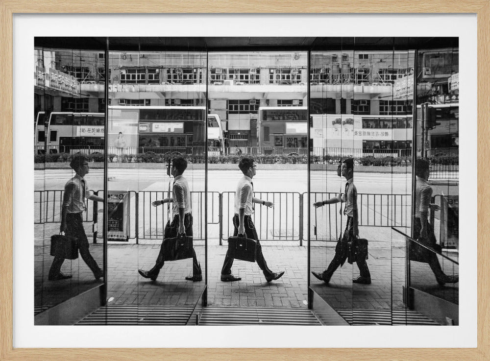 A black and white street photograph showing a man in a shirt and tie carrying a briefcase, his walking motion captured in a time-lapse effect across multiple reflective glass panels. The reflections create a sequence of his stride, set against the backdrop of a busy city street with buses and buildings also reflected in the glass. Decor