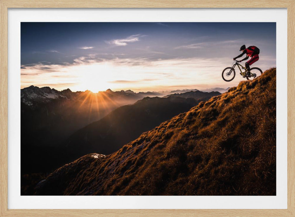 A mountain biker in mid-air, jumping down a steep grassy mountain ridge at sunrise. The sun is low on the horizon, casting a golden glow and long sunbeams over a vast, layered mountain range in the background. Poster