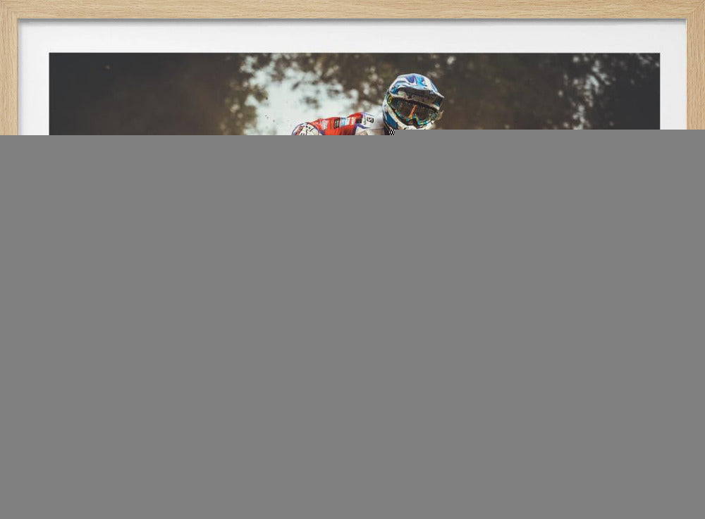 An action shot of a motocross rider in red, white, and blue gear, speeding through a dusty turn on a dirt track, kicking up a large cloud of mud and debris. Another racer is visible in the background. The image is presented within a silver frame. Print