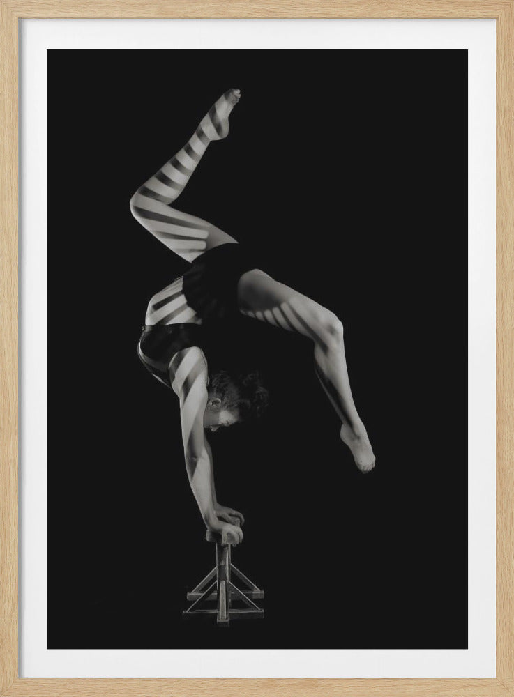 A striking black and white photograph of a female acrobat performing a handstand on a small pedestal. Her body is arched in a deep backbend, and her form is highlighted by dramatic horizontal stripes of light and shadow against a solid black background. Poster