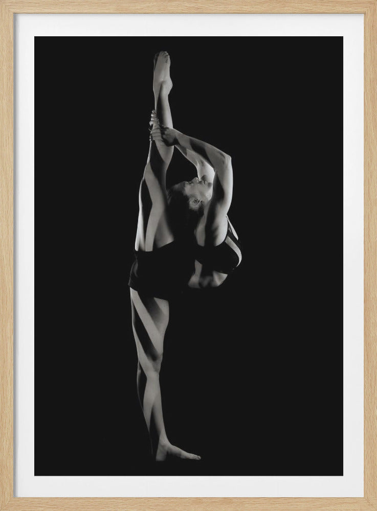 A dramatic black and white studio photograph of a female dancer performing a standing split. She balances on one leg while holding the other leg straight up vertically next to her head, arching her back and looking upwards. The strong side lighting creates a chiaroscuro effect, highlighting her musculature and form against a solid black background. Print