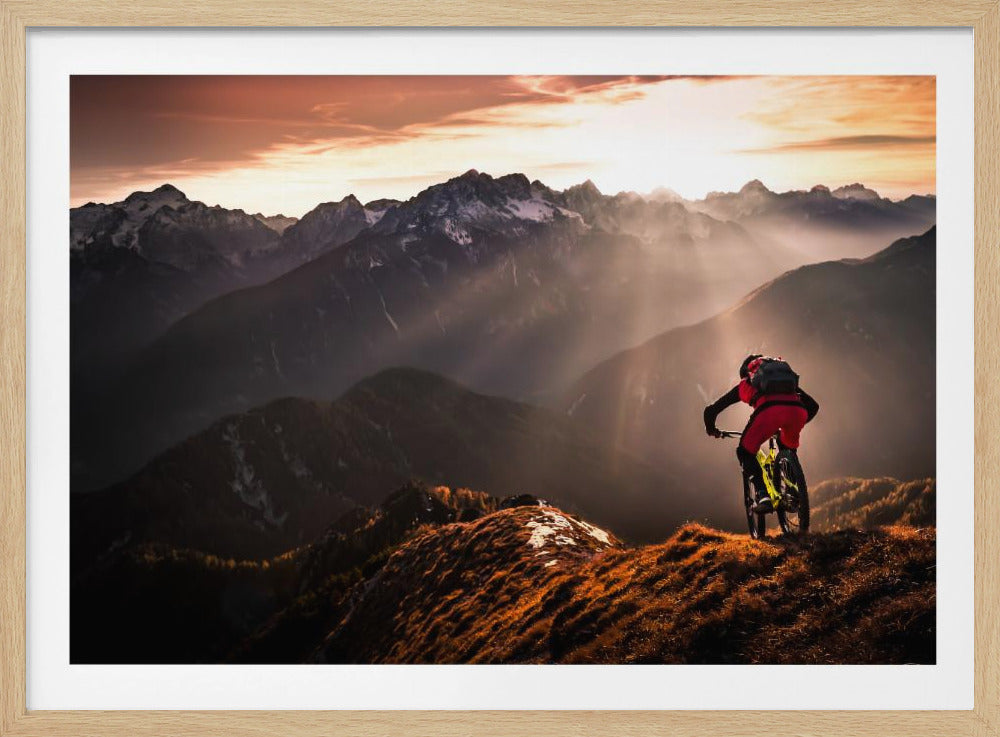 A mountain biker in red and black gear pauses on a grassy, sunlit ridgeline, looking out at a vast mountain range with snow-capped peaks as the sun sets behind them, casting dramatic rays of light through the valleys. Poster