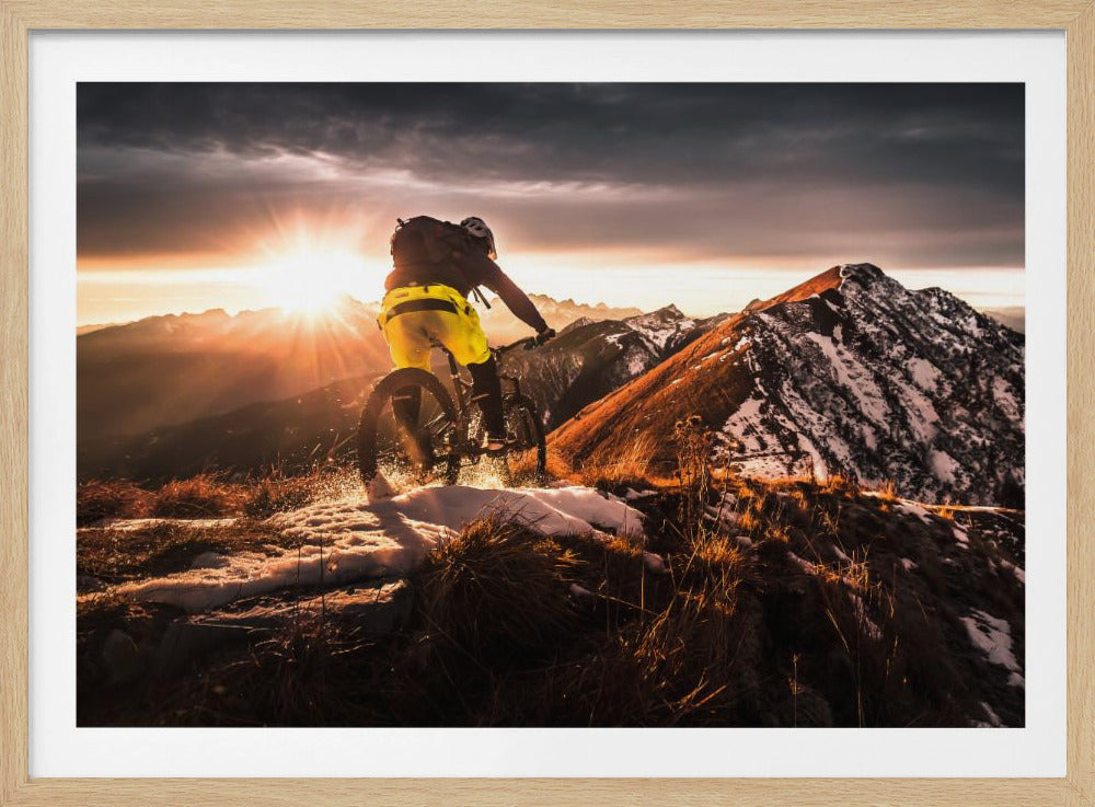 A mountain biker seen from behind rides along a narrow, snowy trail on a mountain ridge during a dramatic sunset, with the sun flaring brightly and a vast, snow-capped mountain range in the background. Decor