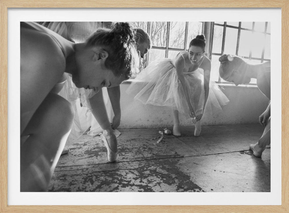 A candid, black and white photograph of several ballerinas in a dance studio preparing for practice. They are all bending over to adjust their pointe shoes by a large, bright window. The entire image is framed by a textured silver border. Wall Art