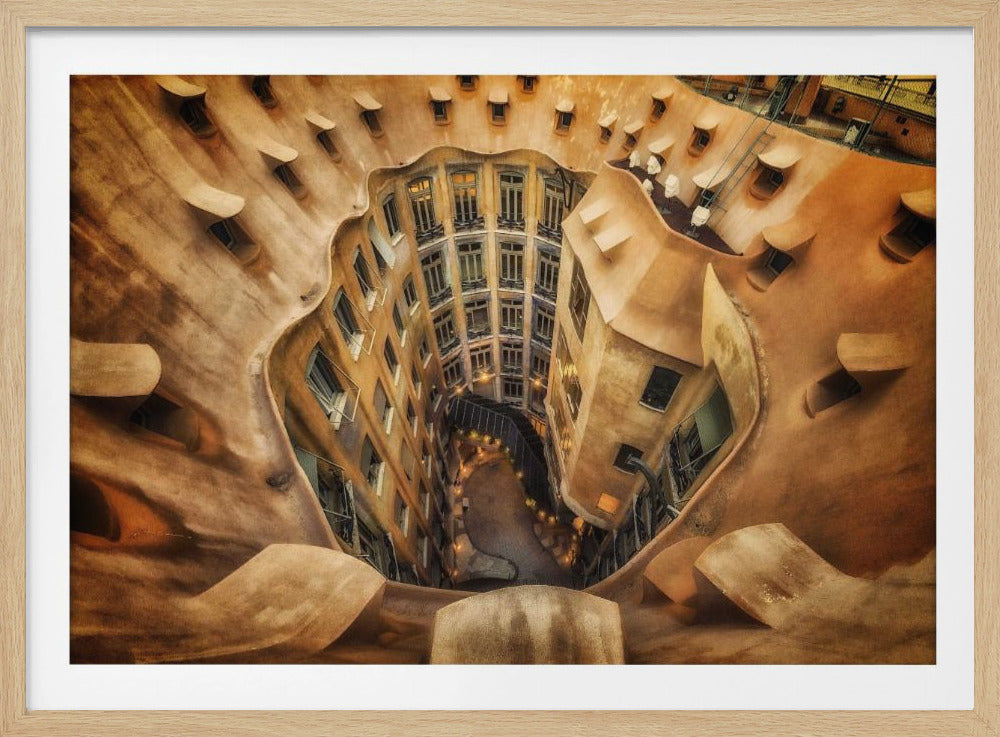 A high-angle, downward view of the inner courtyard of an architecturally unique building, likely Casa Milà by Gaudí. The warm, sandy-colored walls curve and undulate organically, surrounding the multi-story structure with its many windows. The courtyard below is lit, casting a warm glow. Decor