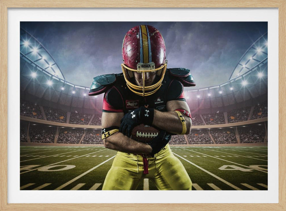 A framed poster of a determined football player in a red and yellow uniform standing on a field in a packed stadium. He is holding a football close to his chest with his head bowed, under bright stadium lights. Decor