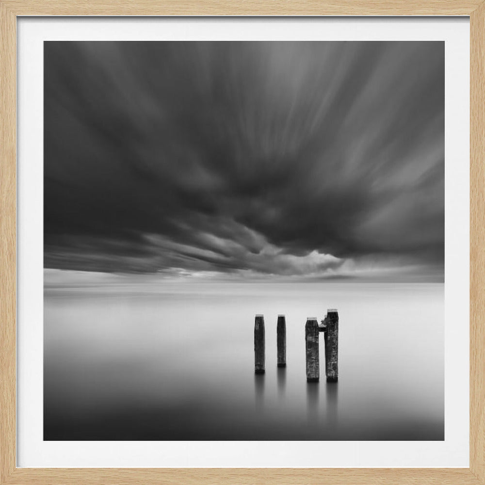 A framed, square, black and white long-exposure photograph of a minimalist seascape. Four dark wooden pilings emerge from still, misty water under a dramatic, streaked sky of dark clouds. Wall Art