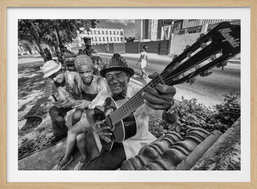 A candid, black and white, wide-angle photograph of three people on a city sidewalk. In the foreground, an elderly Black man in a hat smiles warmly at the viewer while playing a 'Memphis' acoustic guitar. Beside him, a woman in a bandana and another person in a hat also smile, enjoying the moment. The background shows a sunny street with trees and buildings. Decor