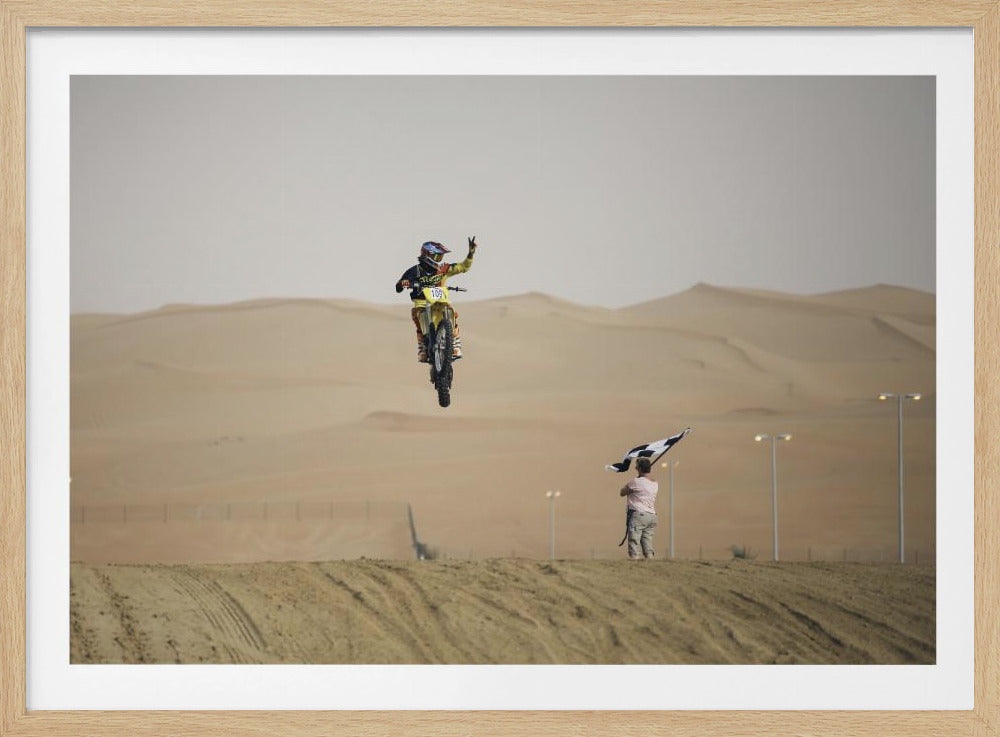 A motocross rider on a yellow dirt bike is captured mid-air during a jump in a desert landscape, flashing a peace sign with their left hand. In the background, rolling sand dunes stretch under a hazy sky, and to the right, a person on a hill waves a black and white checkered flag. Print