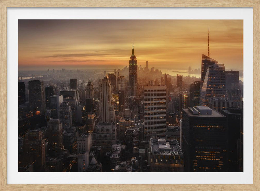 An aerial photograph of the New York City skyline at sunset, with the Empire State Building prominent among numerous skyscrapers under a glowing orange sky, framed in a silver border. Wall Art