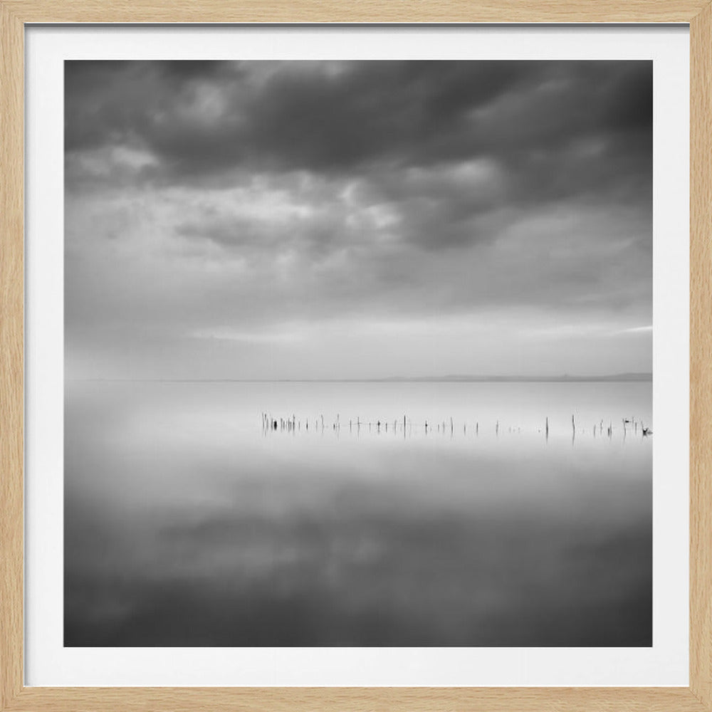 A minimalist black and white photograph in a light wood frame, depicting a calm, serene lake under a heavy, cloudy sky. The still water reflects the sky, with a row of weathered wooden posts breaking the surface along the horizon. Print