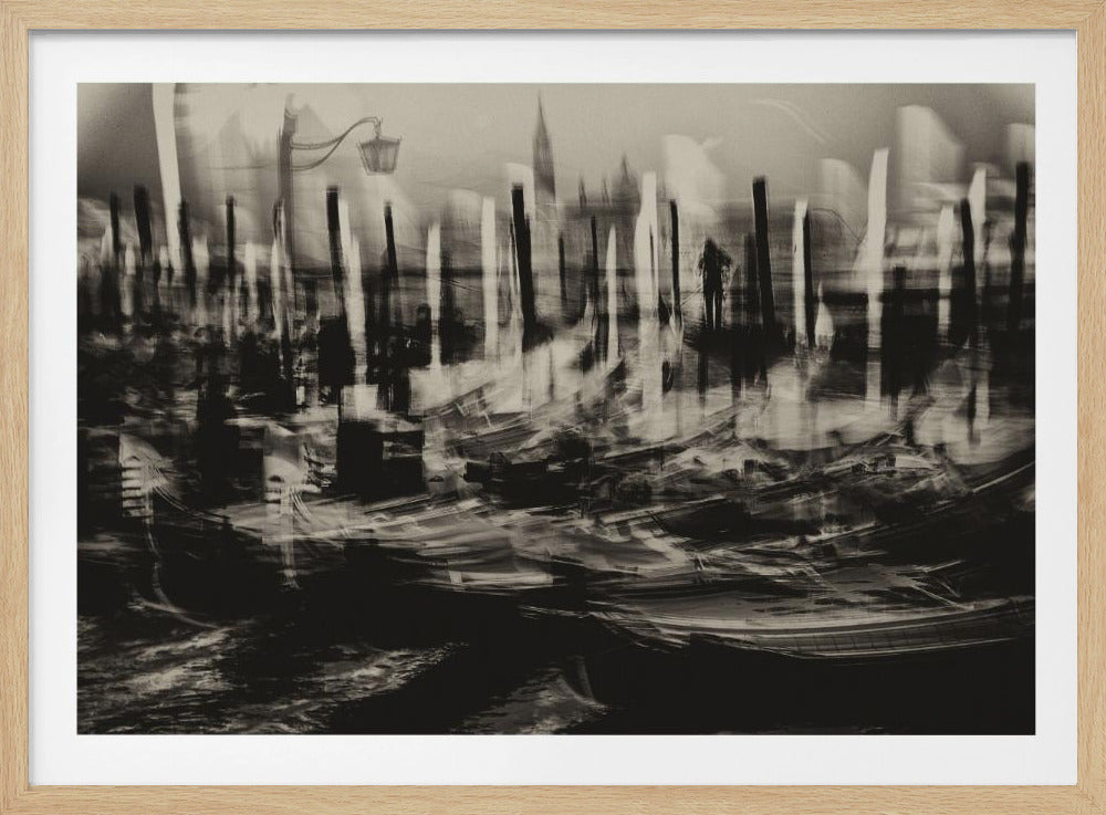 An abstract, black and white photograph capturing the motion of gondolas in a Venetian canal. The image is intentionally blurred, creating streaky, dynamic lines from the boats and their reflections, while mooring poles stand as dark vertical forms against a hazy, indistinct cityscape background. The overall effect is dreamlike and chaotic. Artwork