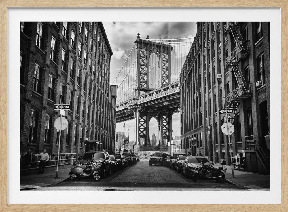 A classic black and white photograph from a cobblestone street in DUMBO, Brooklyn, looking towards the Manhattan Bridge. The bridge is perfectly framed between two large brick buildings. Through an arch of the bridge, the Empire State Building is visible in the distance. The street is lined with parked cars, a scooter, and a motorcycle. The entire image is presented within a silver-colored frame. Print