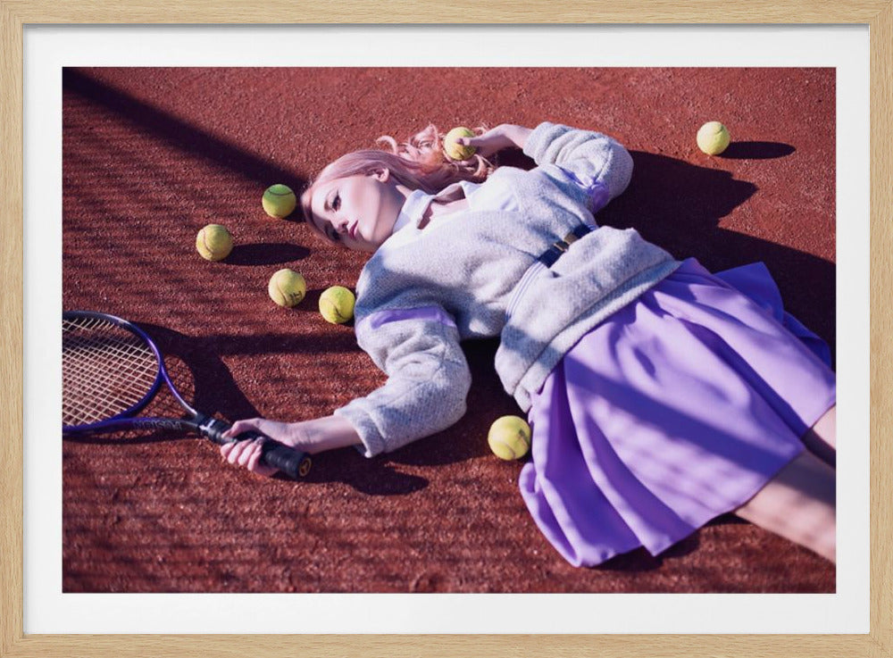A high-angle shot of a young woman with pink hair lying on a red clay tennis court, surrounded by yellow tennis balls. She wears a grey sweater, a white collared shirt, and a purple pleated skirt, and holds a tennis racket in one hand. The scene is framed with a silver border. Wall Art