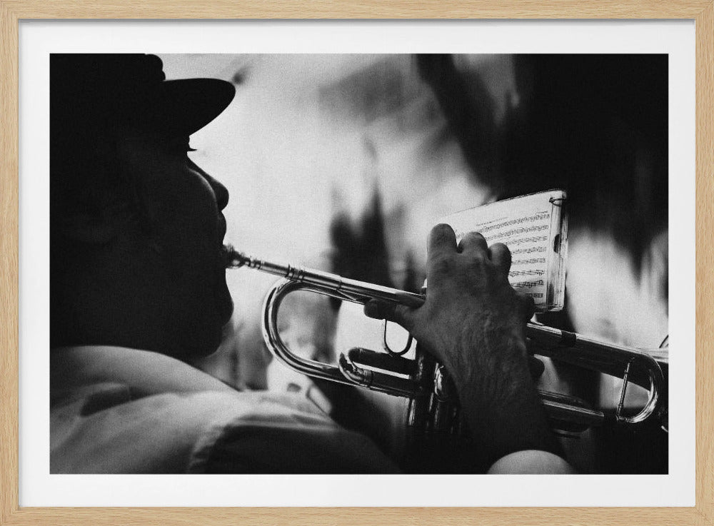 A grainy, black and white close-up photo showing the silhouette of a musician wearing a hat as they play a trumpet. Their hand is visible on the valves, and a small book of sheet music is clipped to the instrument's bell. Print