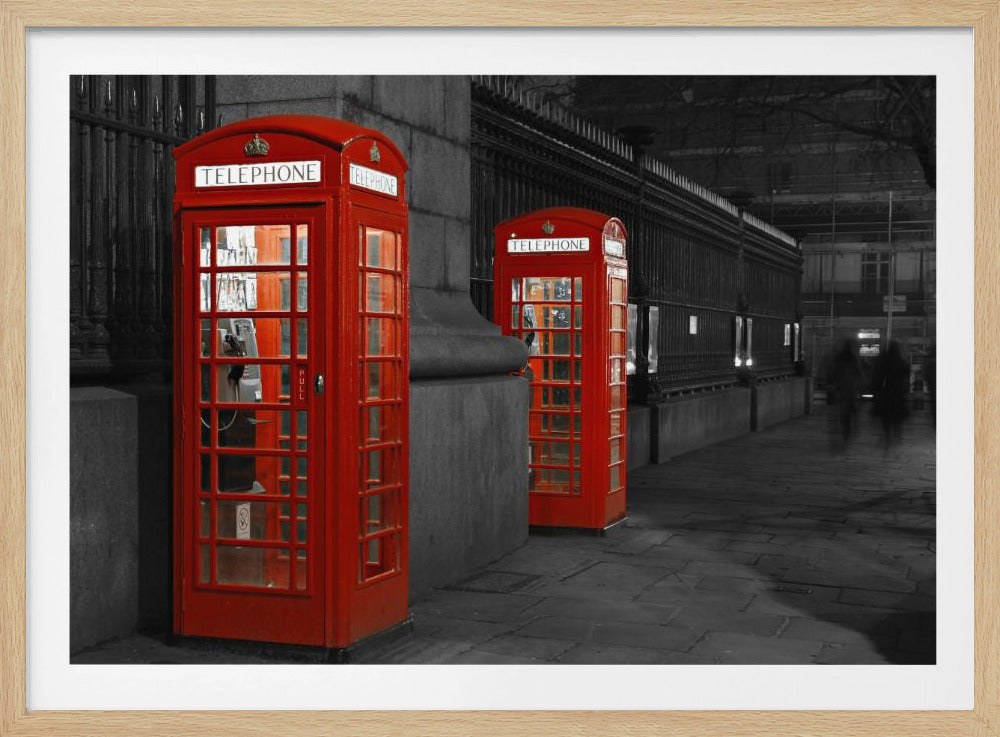 A selective color photograph of a city street at night, framed in silver. Two iconic red telephone booths are the only objects in color, standing out against the black and white background of the cobblestone sidewalk and iron fence. Artwork