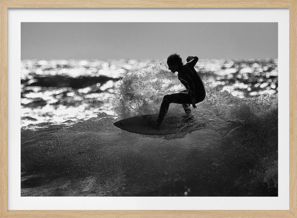 A black and white action shot of a surfer silhouetted against the sparkling ocean. The surfer is crouched low on their board, carving through a wave and creating a dramatic spray of water. The photo is enclosed in a silver frame. Print