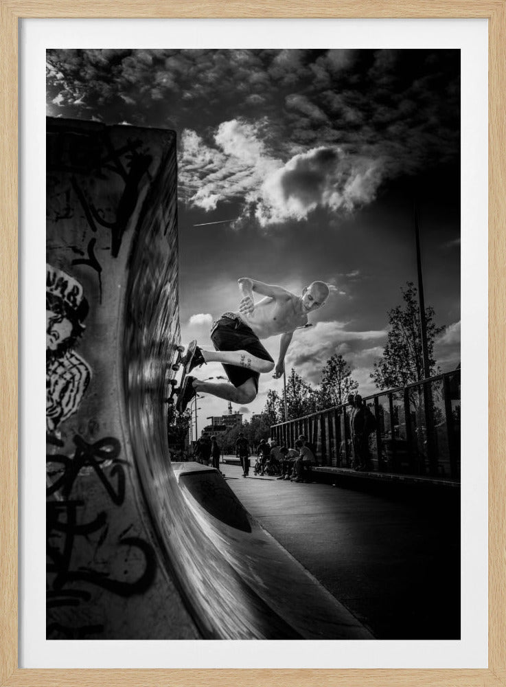 A dramatic black and white, low-angle action shot of a shirtless skateboarder suspended in mid-air at the peak of a graffiti-covered skate ramp, against a backdrop of a cloudy sky. Decor