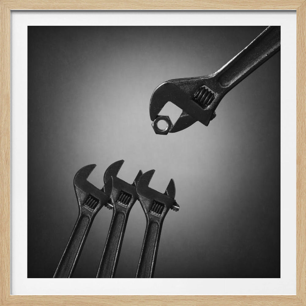 A conceptual black and white photograph in a light wood frame, depicting a large adjustable wrench holding a small nut above three smaller wrenches, which are angled upwards as if they were baby birds waiting to be fed. Print