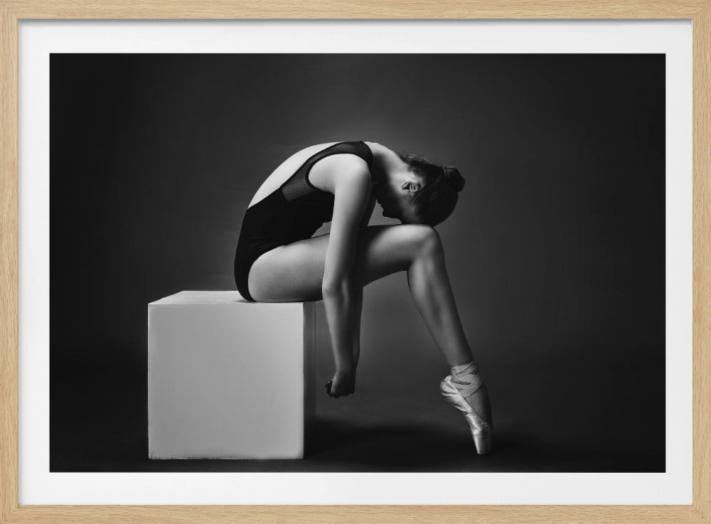 A black and white studio photograph of a ballerina in a contemplative pose, sitting on a white cube against a dark gray background. She is wearing a black leotard and pointe shoes, hunched over with her head resting on her knees. Print
