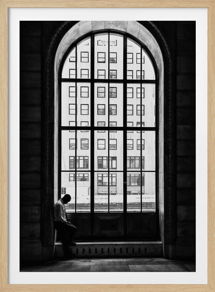 A black and white photograph of a solitary man sitting with his head bowed on the ledge of a massive arched window. The window's dark mullions create a grid over the bright view of another building outside, while the man remains in the shadows, creating a pensive and lonely mood. Poster