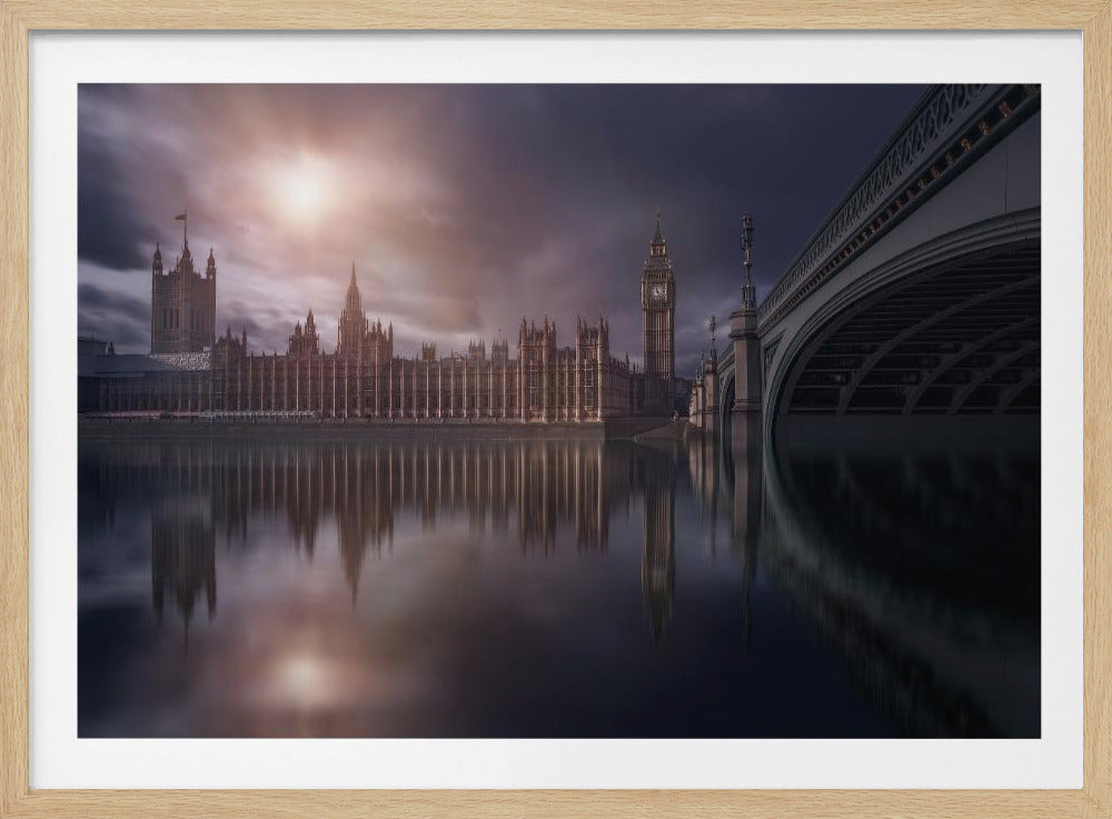 A dramatic, atmospheric photograph of the Houses of Parliament and Big Ben in London, reflected in the still waters of the River Thames. A bright sun breaks through dark, moody clouds, casting a warm light on the scene, with a stone bridge visible to the right. The image is enclosed in a silver frame. Print