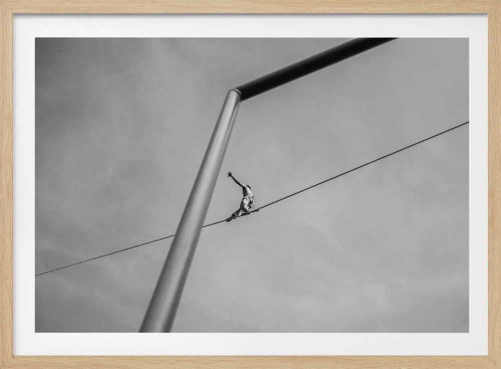 A black and white, low-angle photograph of a person balancing while sitting on a tightrope high in the sky. A large metal pole is in the foreground, creating a strong diagonal line. The image is framed in silver. Decor