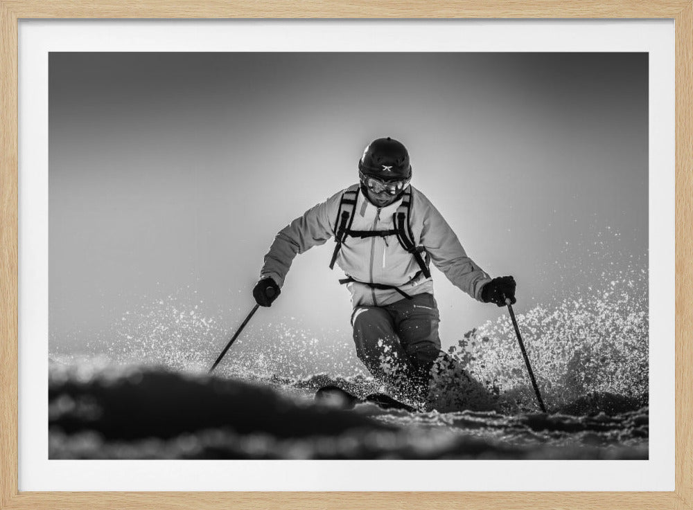 A black and white action photograph of a skier in full gear, leaning into a turn and spraying powder snow. The skier is backlit by the sun, creating a high-contrast, dramatic effect. Print