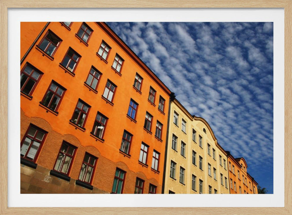 A low-angle shot of colorful buildings, predominantly a vibrant orange one, against a bright blue sky filled with a pattern of white, fluffy clouds. The image is presented within a silver-colored frame. Print