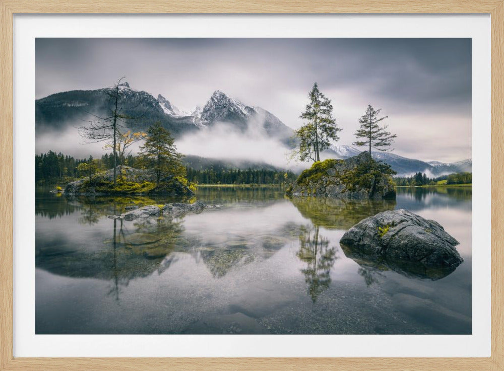 A serene and misty mountain lake scene with several small rocky islands topped with evergreen trees. In the background, snow-capped mountains rise through a layer of fog under a cloudy gray sky. The calm, clear water reflects the mountains and islands perfectly. Print