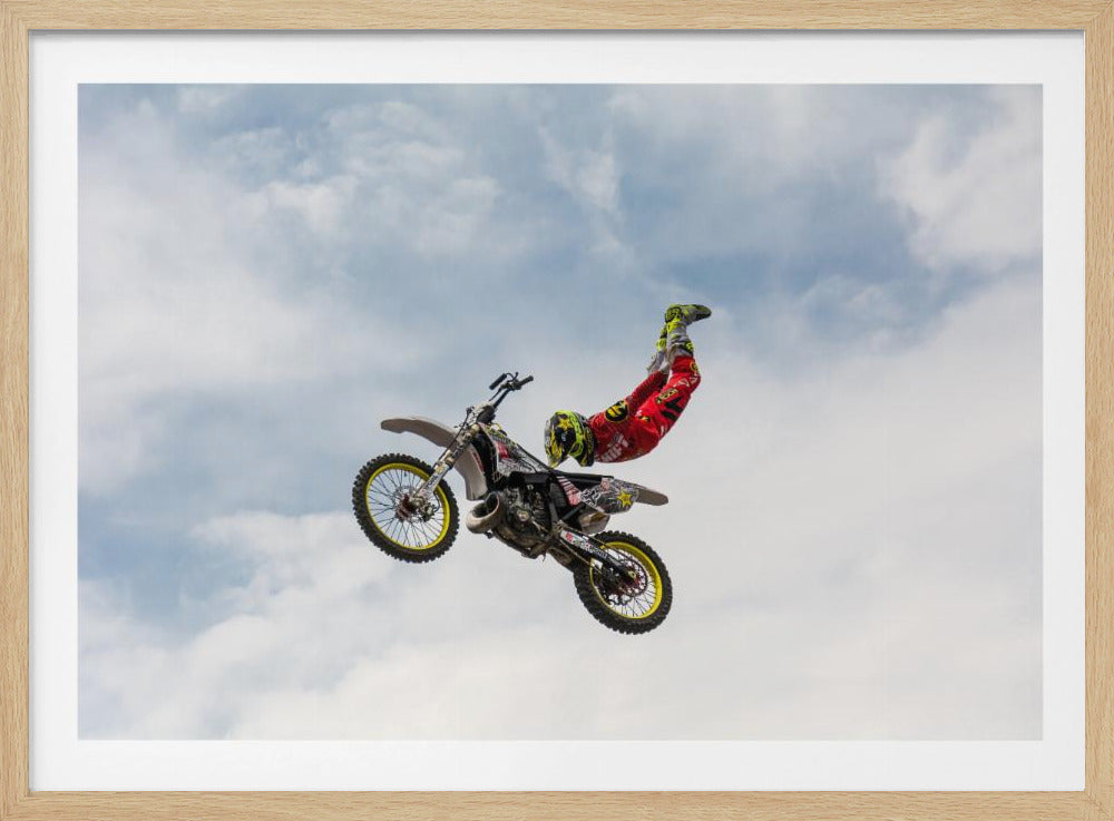 A motocross rider in red and yellow gear performs a high-flying stunt, arching their back while upside down in the air next to their dirt bike, set against a backdrop of a blue sky with white clouds and enclosed in a silver frame. Poster