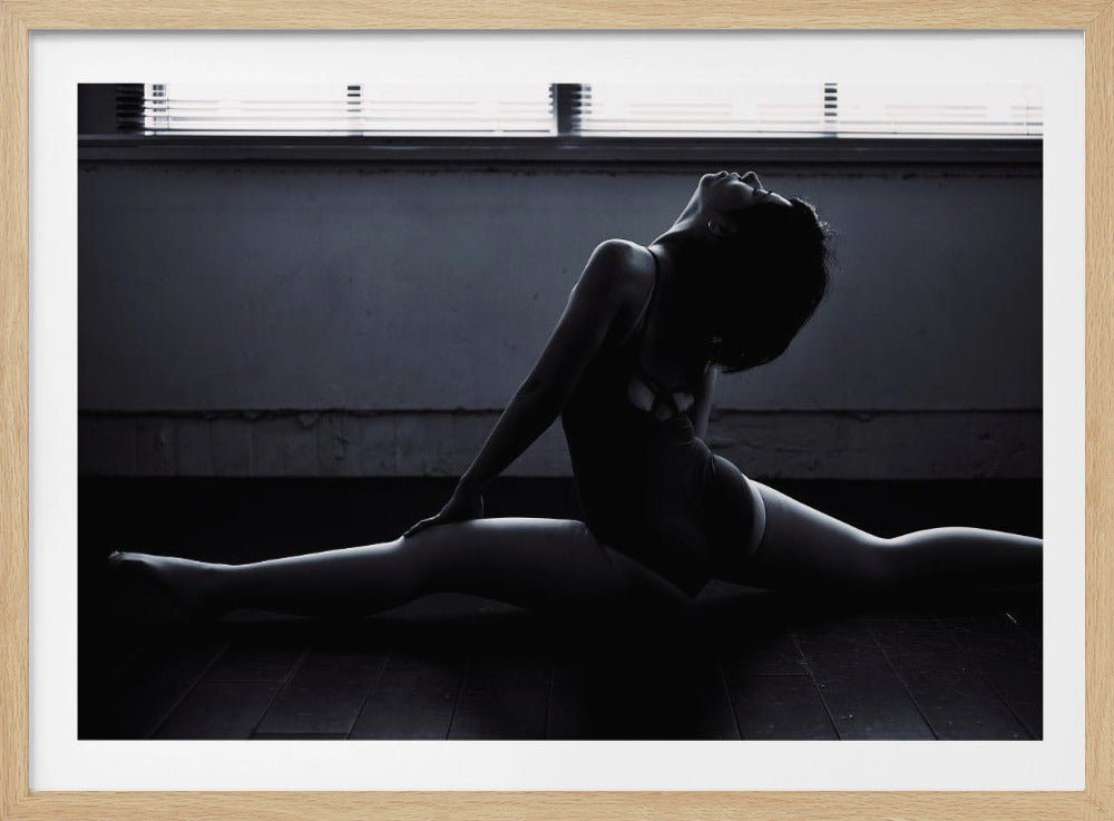 A dramatic, low-key black and white photograph of a female dancer with short hair in a leotard, holding a full split on a dark wooden floor. Her back is arched and her head is tilted up, with side lighting creating strong highlights and shadows on her form. The image is presented in a silver frame. Decor
