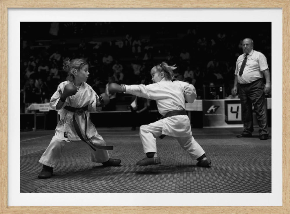 A black and white photograph of two young girls in a karate competition. They are wearing white gis, black belts, and boxing gloves. One girl is in the middle of throwing a punch at her opponent. In the background, a referee stands watching the match, and spectators are blurred in the stands. The image is presented within a silver-colored frame. Print