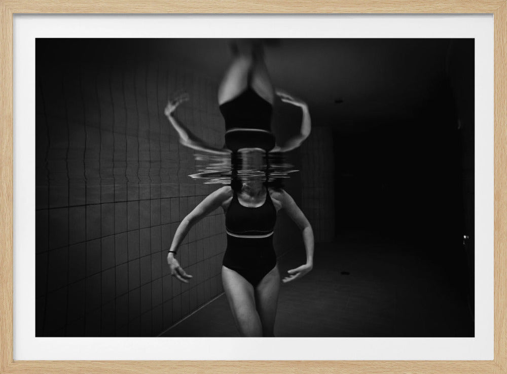 A dramatic black and white photograph of a woman in a one-piece swimsuit in a tiled pool. Her upper body is submerged, while her lower body is perfectly reflected on the water's surface above her, creating a surreal and symmetrical image. Wall Art