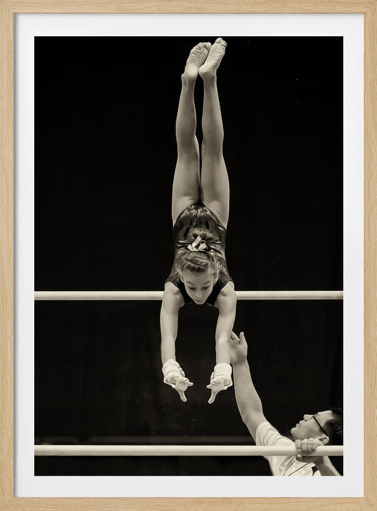 A dramatic black and white photograph of a young female gymnast performing a flawless handstand on the top of two parallel bars. Her body is perfectly vertical against a stark black background. Below, a male coach looks up at her with focused attention, his hand raised nearby, ready to spot her. Decor