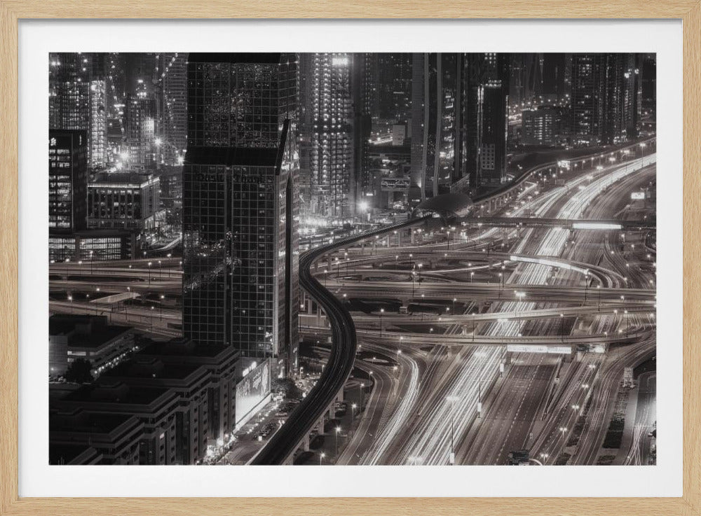 A dynamic black and white long-exposure photograph of a sprawling metropolis at night, viewed from above. Gleaming skyscrapers fill the background, while a complex interchange of highways with streaks of light from traffic dominates the foreground, with an elevated train line weaving through the city. Wall Art