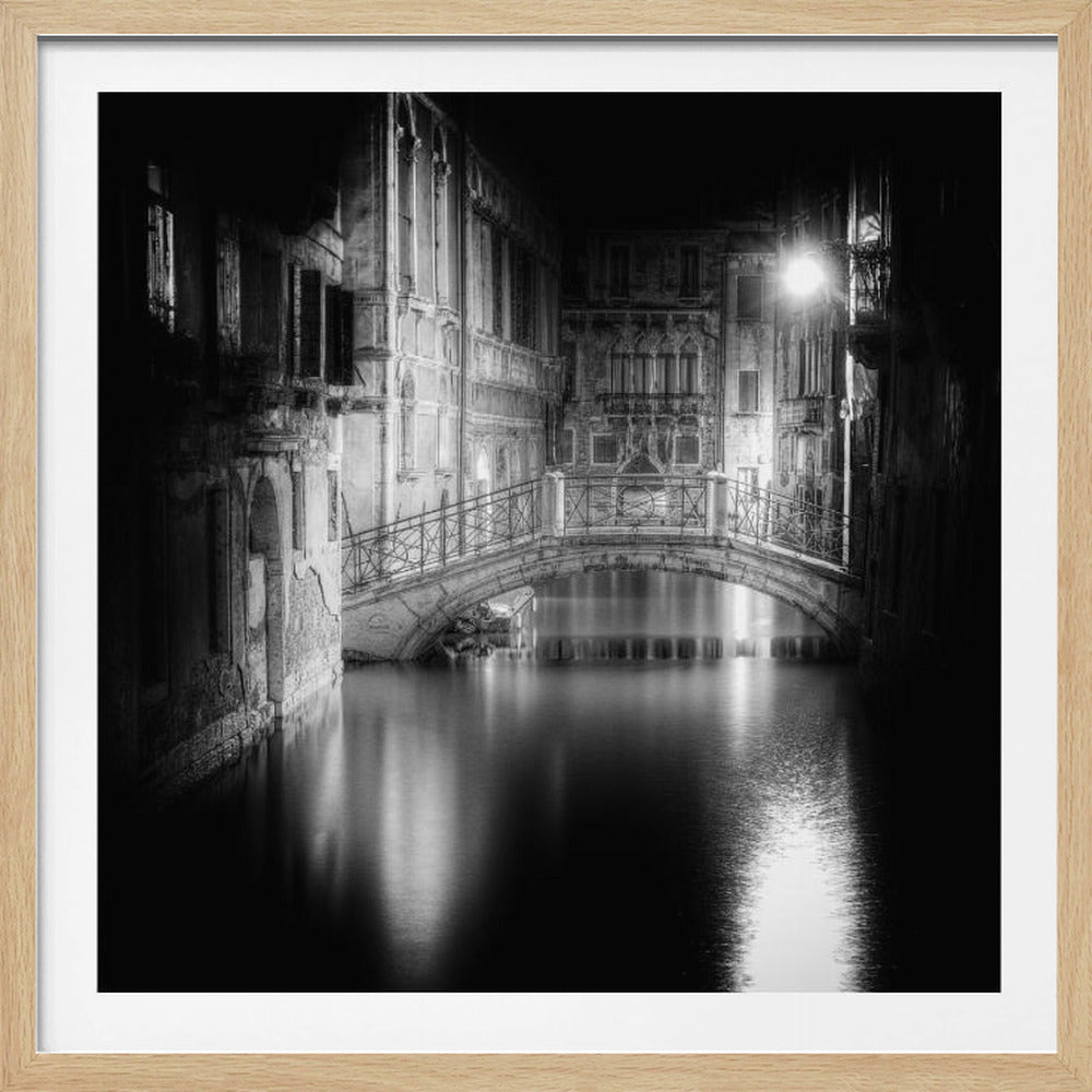 A dramatic black and white long-exposure photograph of a Venetian canal at night. An ornate, arched footbridge crosses the smooth, dark water, which reflects the light from a single bright lamp and the illuminated historic buildings lining the canal. Artwork