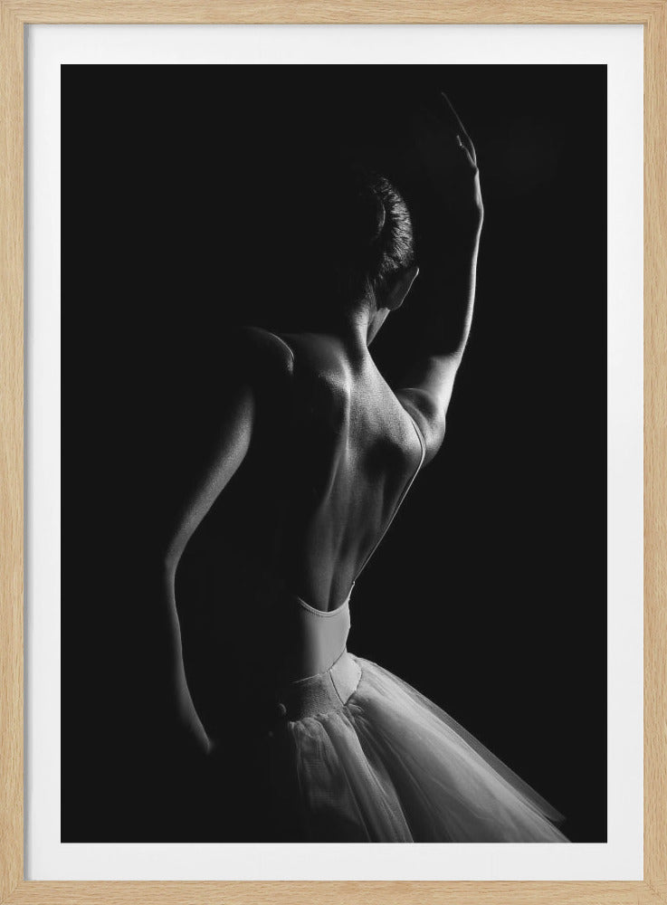 A dramatic black and white photograph of a ballerina viewed from behind against a solid black background. The lighting from the right side highlights the defined muscles of her back, her raised arm, and the edge of her white tutu, creating a strong contrast between light and shadow. Print